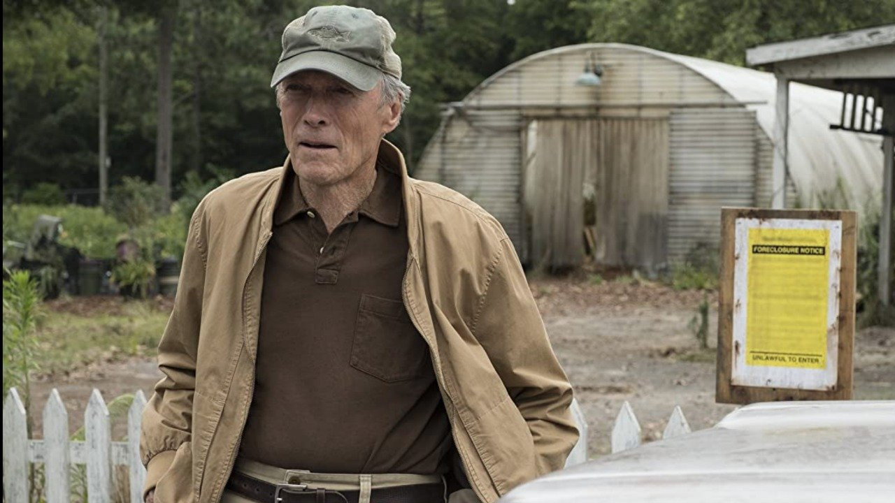 Vemos Earl Stone, interpretado por Clint Eastwood no filme A Mula. Ele é um homem branco, idoso, de boné cinza, jaqueta bege e camiseta marrom.