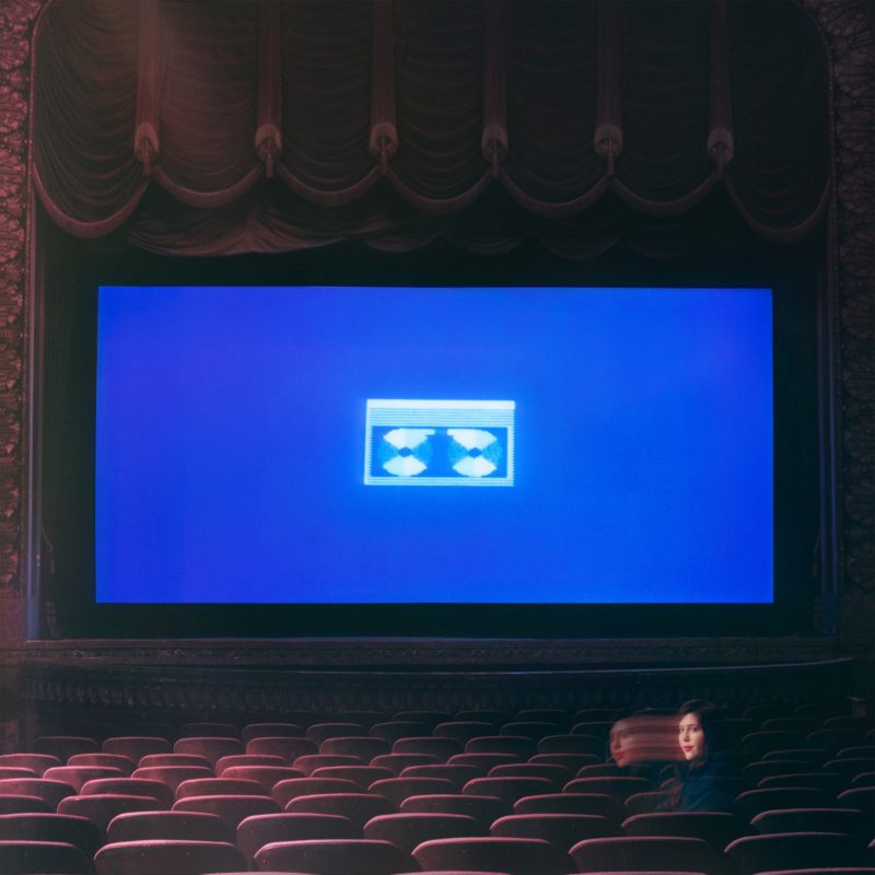  Capa do álbum Home Video, da cantora Lucy Dacus. Uma sala de cinema quase vazia, com poltronas vermelhas e uma cortina levantada. Apenas Lucy está presente, sentada na parte direita da sala, com seu rosto em movimento, criando um espectro de sua face à esquerda. A tela está coberta por uma projeção azul, com o ícone de uma fita cassete no centro.