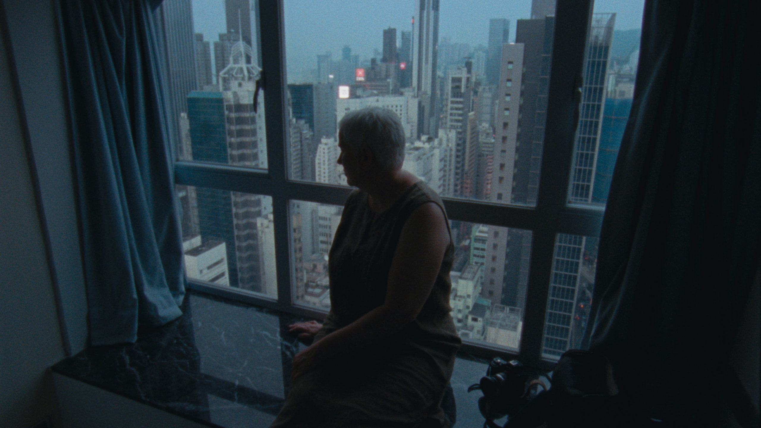 Cena do filme Madeira e Água. A imagem mostra uma mulher branca de cabelos bem curtos e brancos, sentada ao lado de uma grande janela através da qual vemos inúmeros prédios de uma grande cidade. 