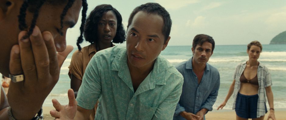 Cena do filme Tempo exibe pessoas numa praia acudindo um homem negro, com tranças no cabelo, que está com o nariz sangrando. As outras pessoas parecem apreensivas. 