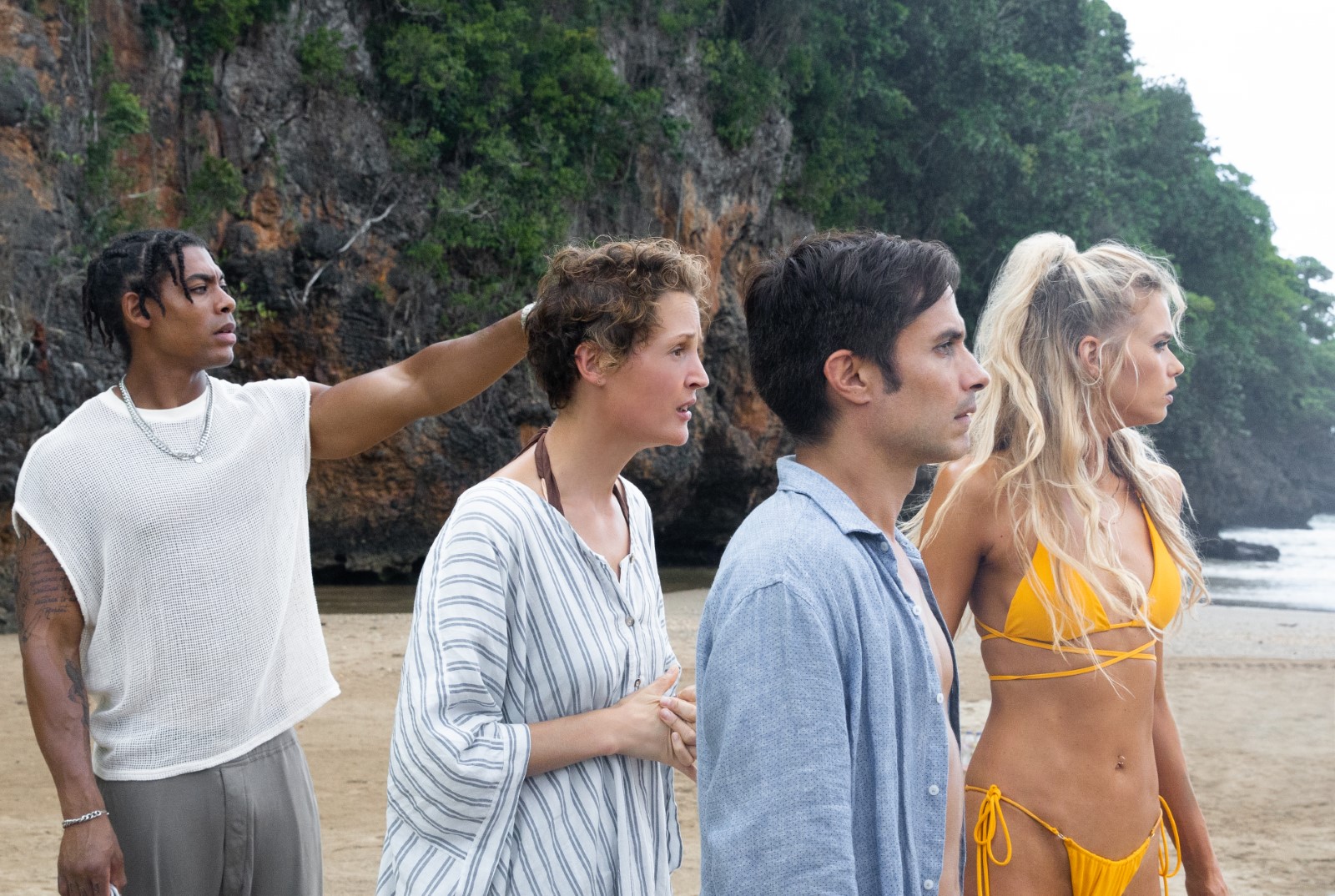  Cena do filme Tempo exibe quatro pessoas numa praia olhando para o mar. Um homem negro aponta para o oceano. Um homem branco e duas mulheres também brancas observam as ondas. 