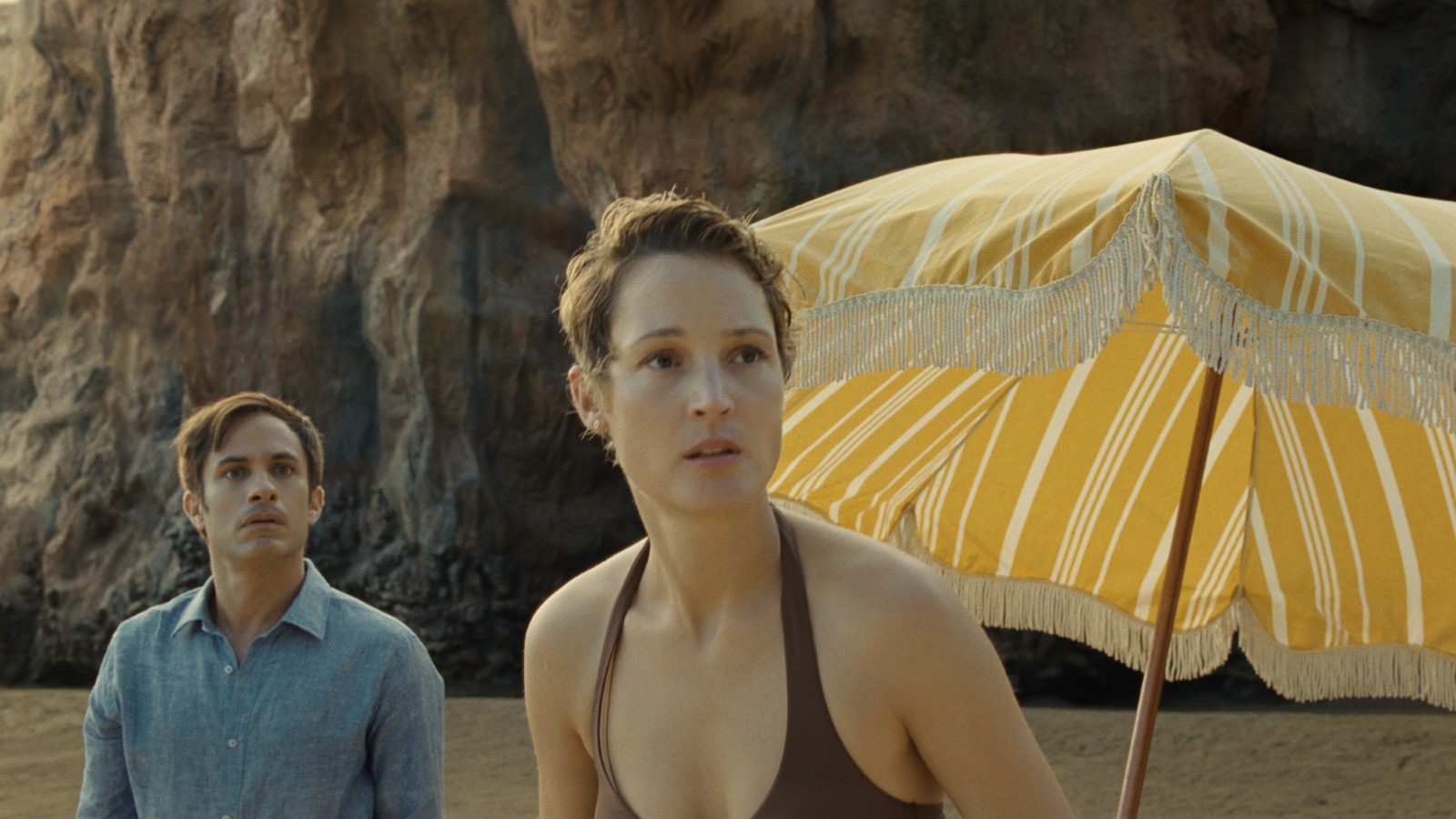 Cena do filme Tempo exibe um casal numa praia. Ambos são brancos. O homem veste uma camisa azul e a mulher usa um biquíni marrom. No lado direito, vemos um guarda-sol amarelo. 