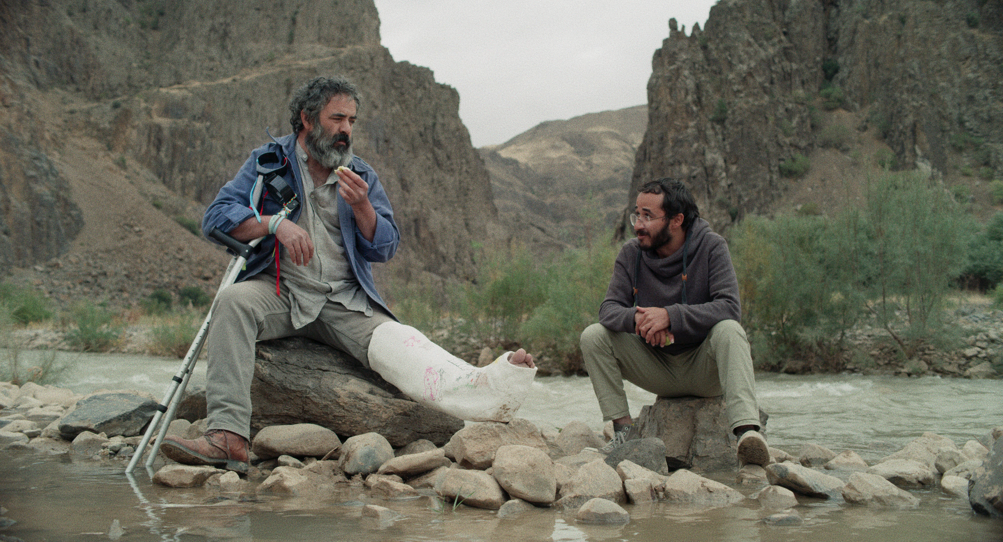 Cena do filme Pegando a Estrada. A foto mostra dois homens persas sentados sobre pedras no meio de um riacho. O da esquerda é mais velho, tem barba grisalha, a perna esquerda engessada e duas muletas apoiadas em seu corpo. O outro é jovem, tem óculos, cabelos escuros e barba.