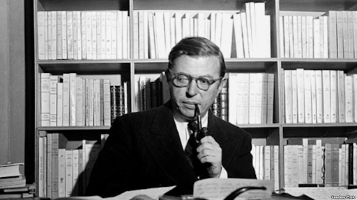 Foto em preto e branco do filósofo Jean-Paul Sartre, na foto ele segura um cachimbo e o seu fundo é composto por vários livros.