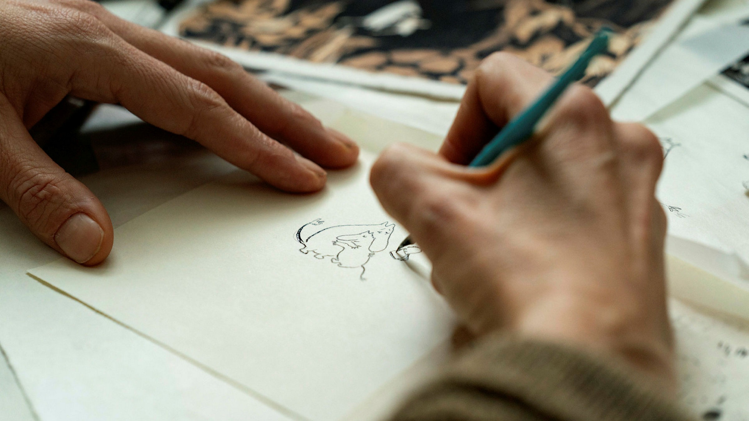 Cena do filme Tove. Na imagem, vemos, em close up, duas mãos brancas desenhando em um papel. A mão direita segura um lápis azul e desenha dois personagens Moomins, criaturas fictícias que lembram hipopótamos.