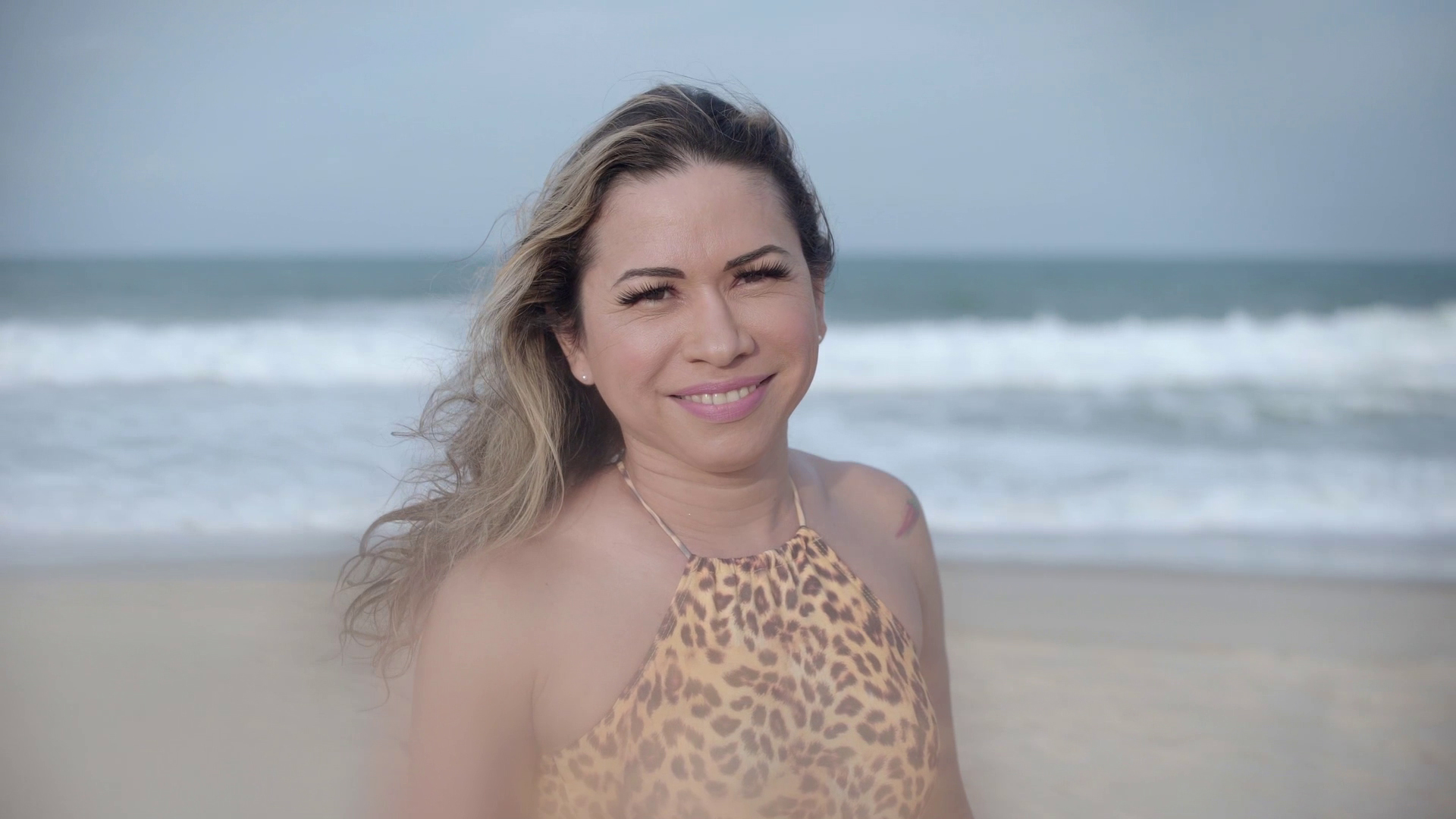 Cena do documentário Transversais mostra uma mulher loira sorrindo na praia.