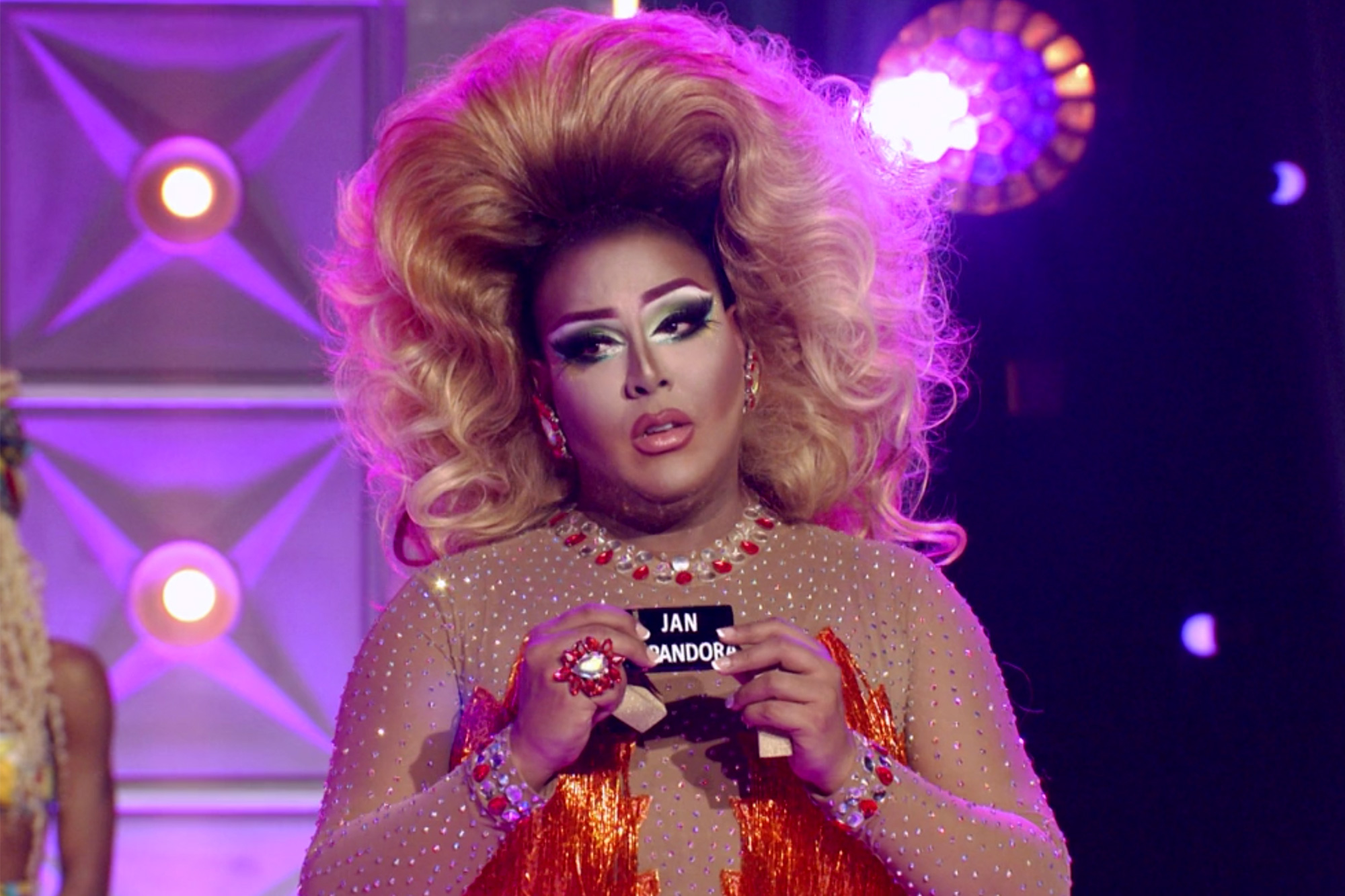 Cena do reality show All Stars 6, mostra a drag queen latina Alexis Mateo, de vestido laranja e peruca loira, segurando dois batons. As embalagens são pretos e, em branco, lemos Jan e Pandora em cada um deles.