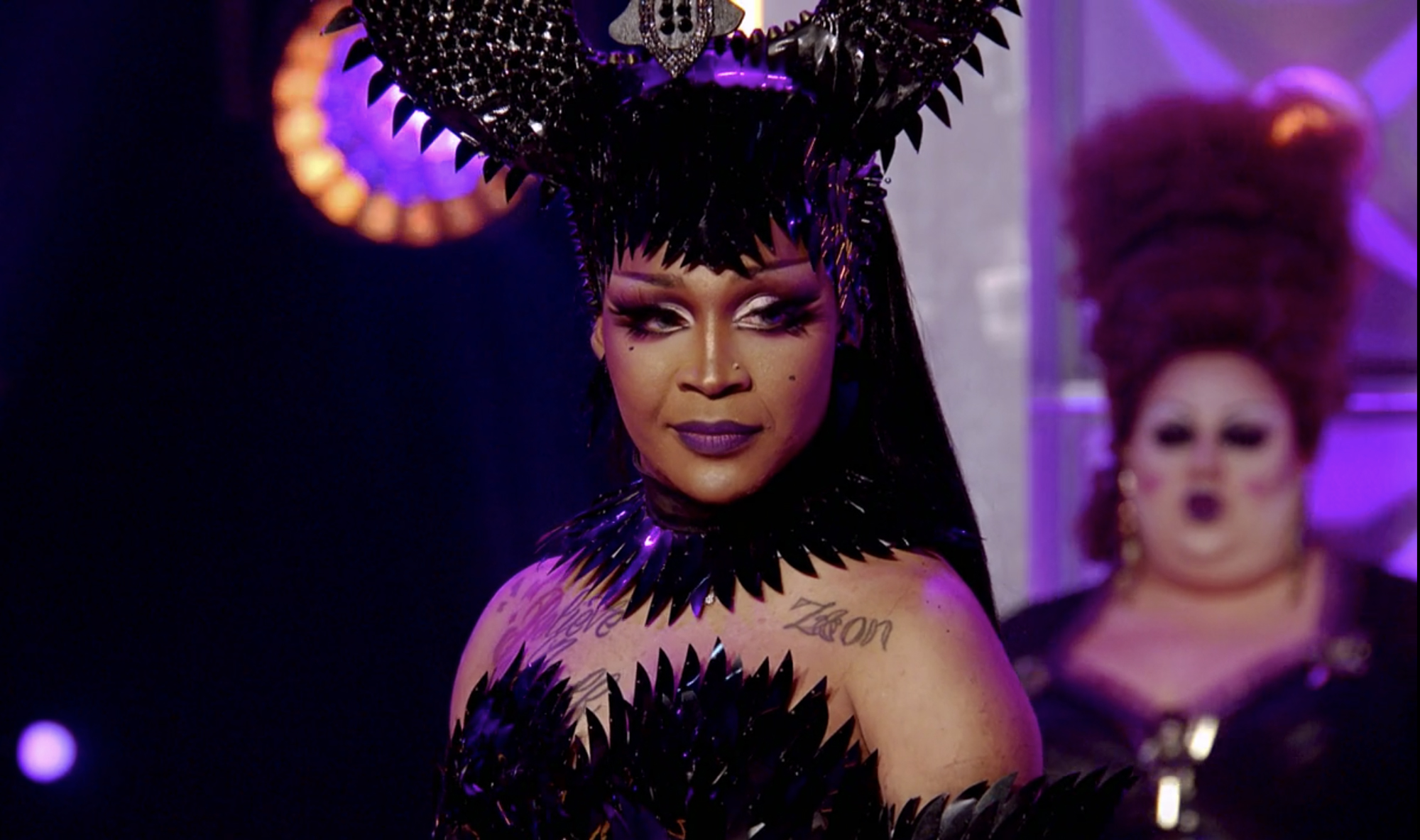 Cena do reality show All Stars 6, mostra o semblante triste de A'keria C. Davenport, drag queen negra, que usa vestido e ornamento na cabeça pretos e pontiagudos, na passarela.