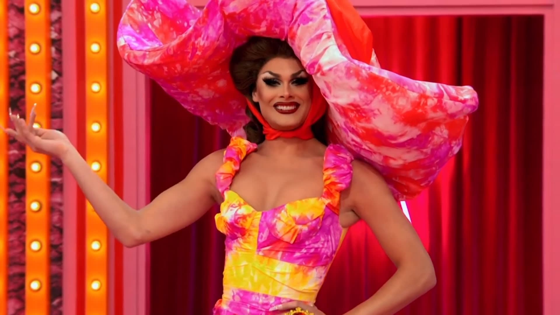 Cena do reality show All Stars 6, mostra a entrada da drag queen branca e magra Scarlet Envy. Ela sorri, e veste vestido e chapelão em tie-dye, misturando as cores amarelo, rosa e roxo. Ao fundo, vemos as cortinas vermelhas do Ateliê.