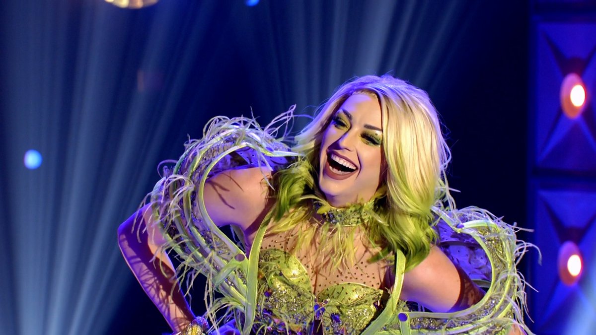 Cena do reality show All Stars 6, mostra a drag queen branca e loira Laganja Estranja sorrindo; ela usa roupas verdes e se enverga para frente, sorrindo de boca aberta e com os dentes a mostra.