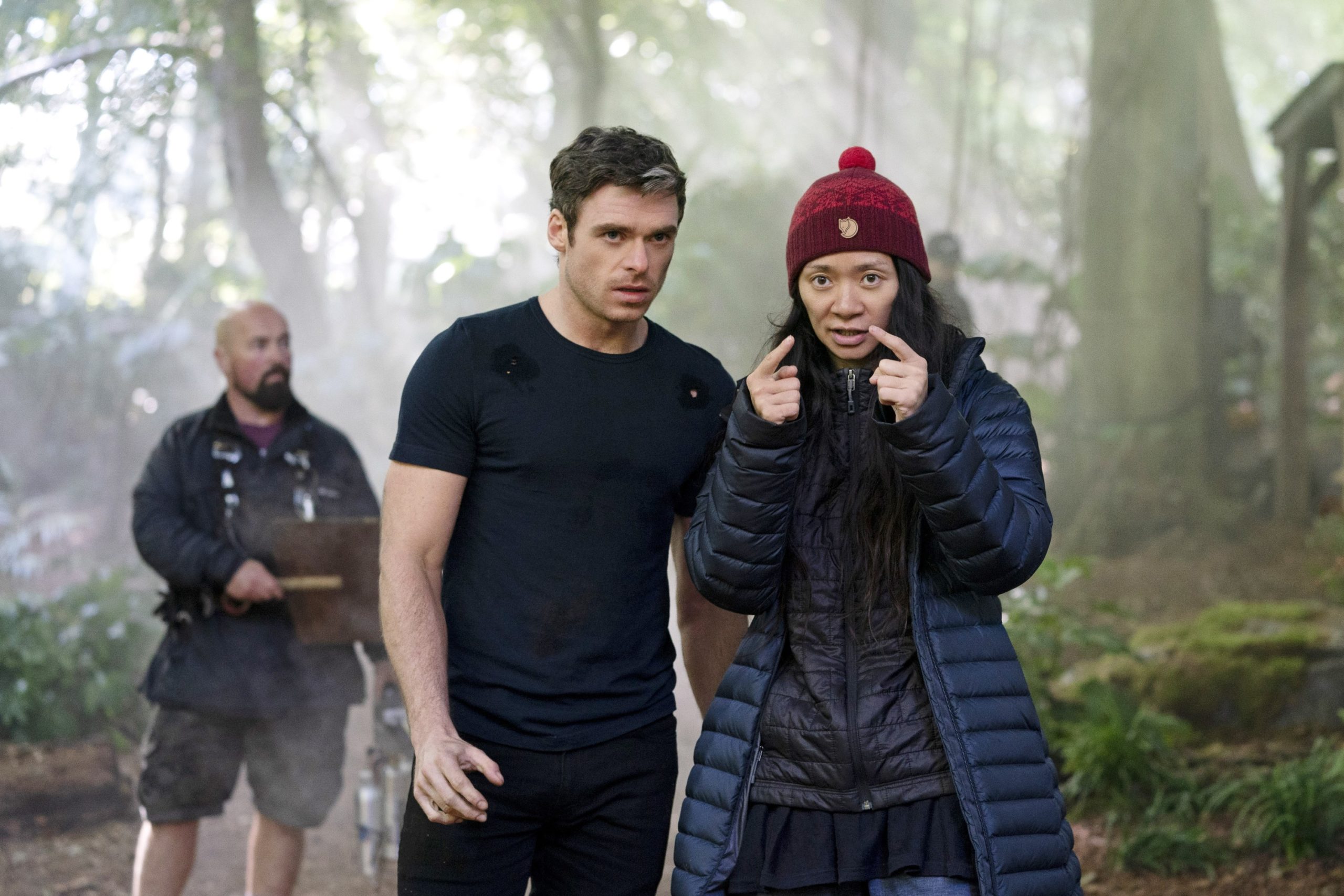Foto dos bastidores do filme Eternos. A foto é em uma floresta, e a diretora Chloe Zhao passa instruções para o ator Richard Madden. Ela veste um gorro vermelho e casaco azul, enquanto ele usa uma camiseta e calças pretas.