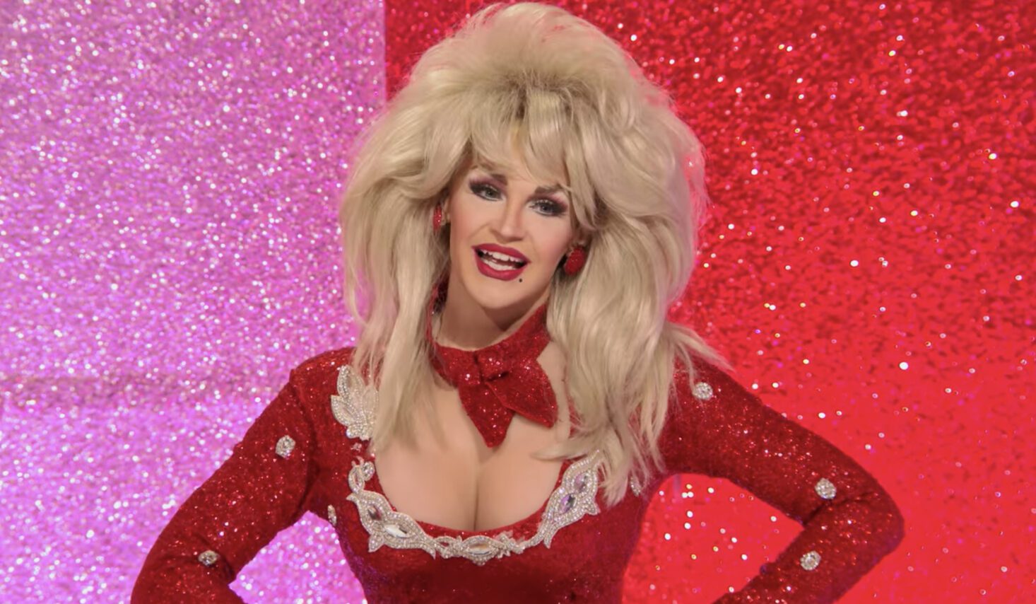 Cena do reality show All Stars 6. Na foto, vemos a drag queen Kylie Sonique Love imitando a cantora Dolly Parton. Ela sorri, usa uma peruca loira bem volumosa e um vestido vermelho com decote avantajado. 