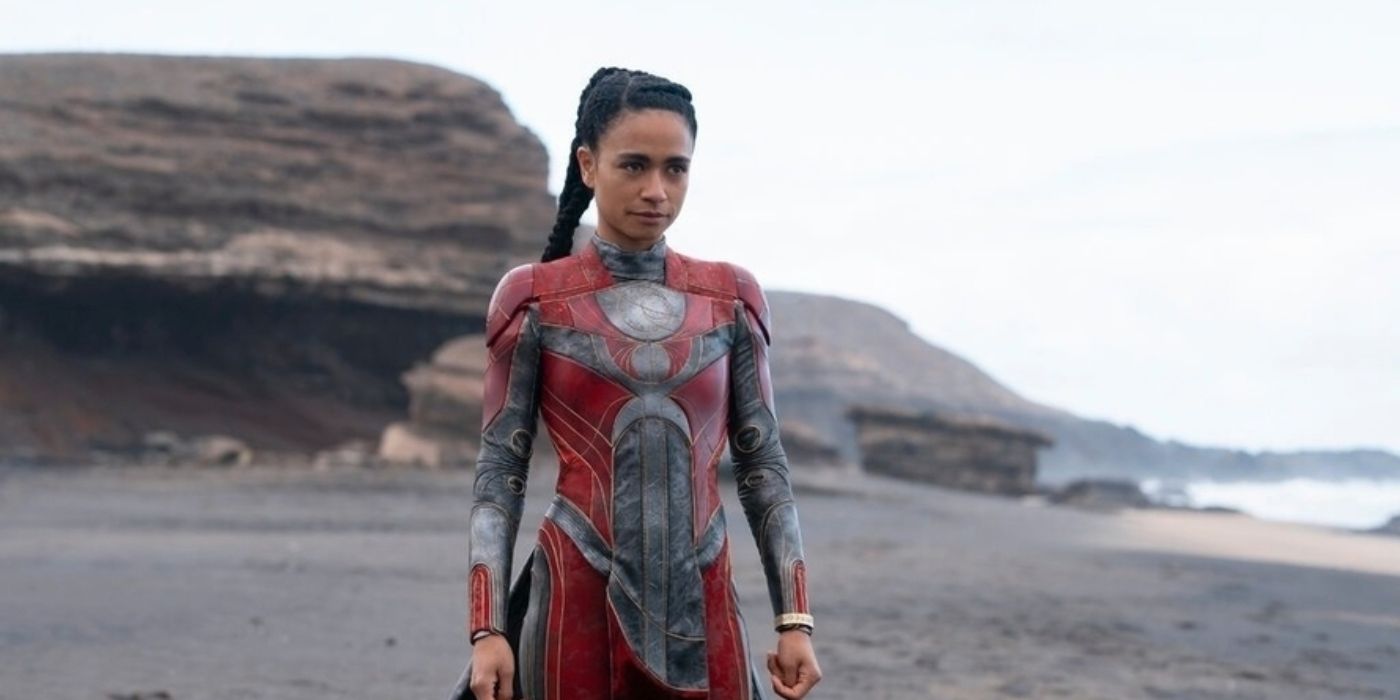 Cena do filme Eternos, mostra Makkari, uma mulher negra e que usa uma armadura vermelha, olhando para frente, e o cenário é uma praia à luz do dia.