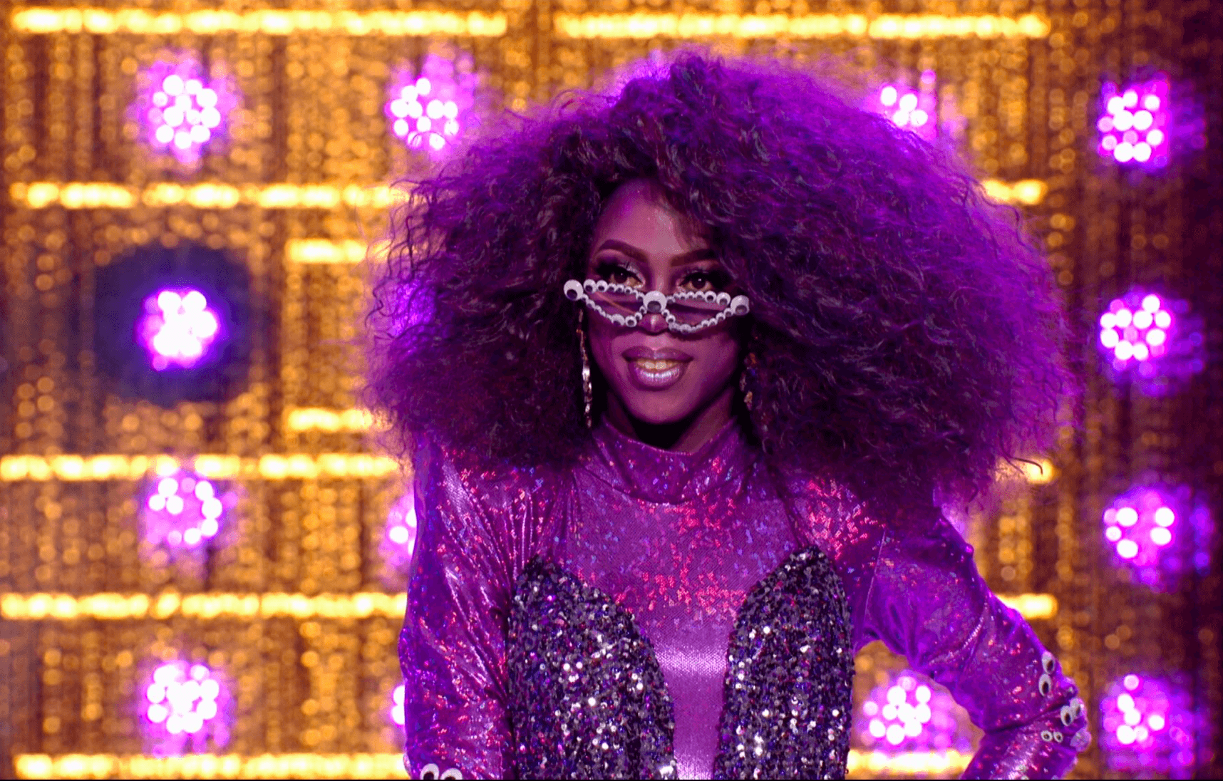 Cena do reality show All Stars 6, mostra Ra'Jah O'Hara na passarela, vestindo um macacão roxo, com o rosto, a peruca e os óculos pintados da mesma cor. Ela sorri e olha por cima dos óculos escuros.