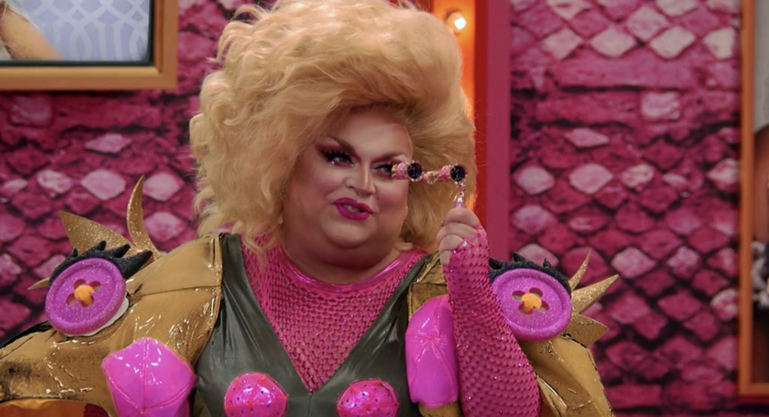 Cena do reality All Stars 6, mostra Ginger Minj, uma drag queen branca e gorda, vestida de sapo, segurando uma espécie de binóculos na frente do rosto e sorrido, se dirigindo à alguém que está na frente dela.