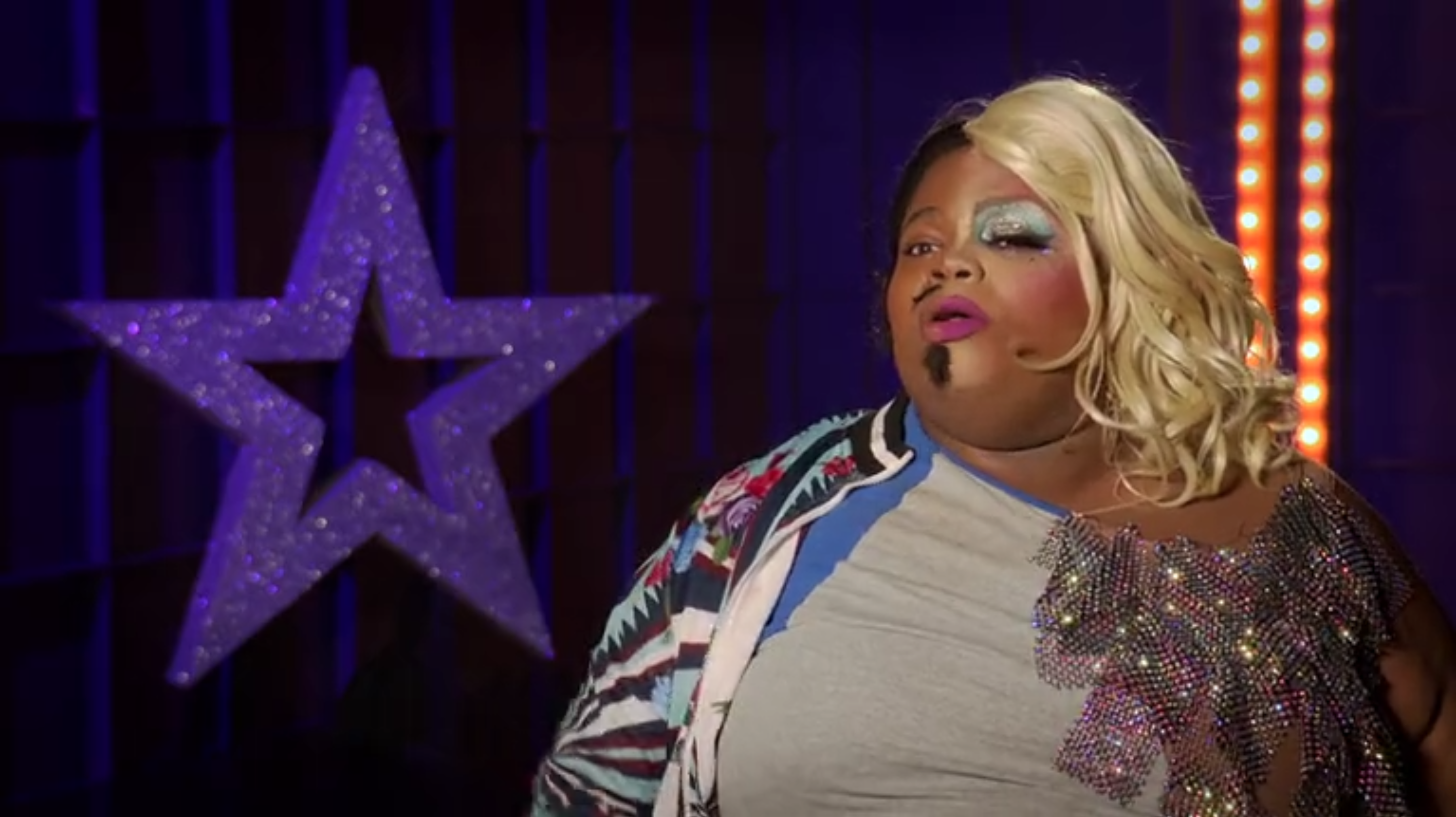 Cena do reality All Stars 6, mostra Silky Nutmeg Ganache dando entrevista. Ela é uma drag queen negra e gorda, que está vestida meio a meio. No lado direito, tem a parte masculina, com bigode e barbicha, e uma roupa azul, enquanto o lado esquerdo é o feminino, com vestido e peruca loira.