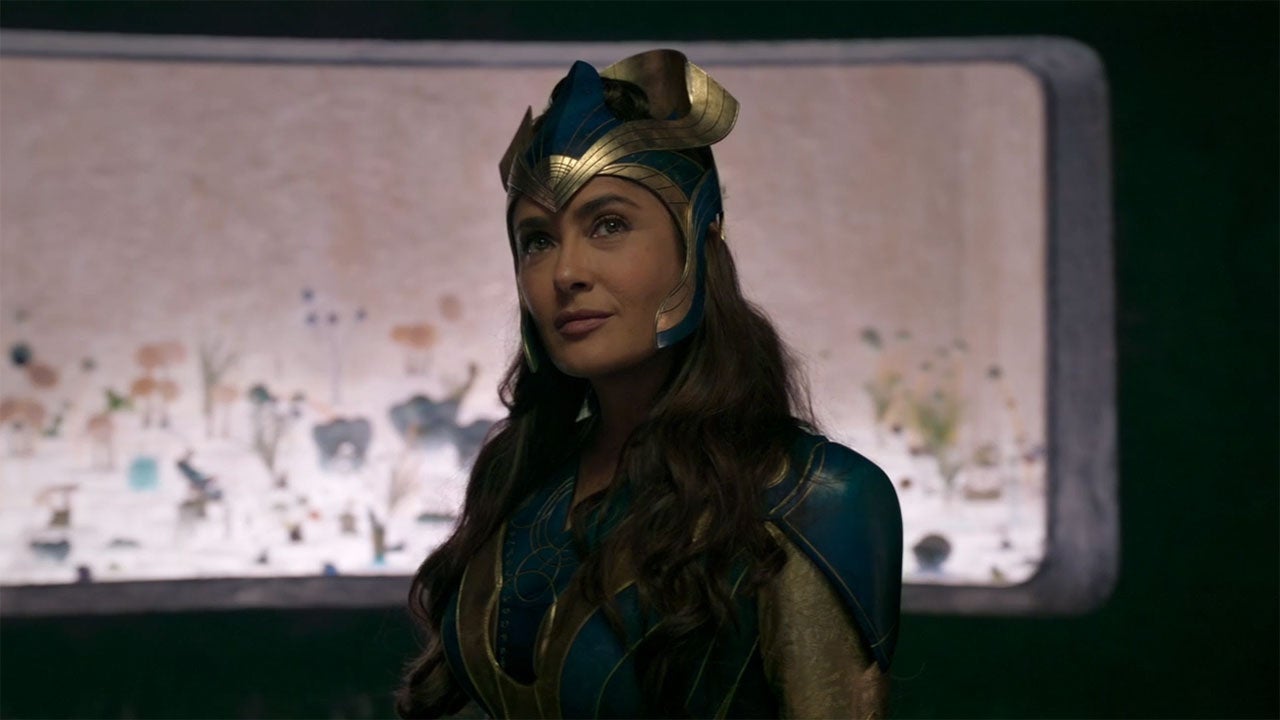 Cena do filme Eternos, mostra Salma Hayek sorrindo. Ela é uma mulher latina, de cabelos castanhos e veste uma armadura verde, com direito a ornamento na cabeça, em tons de dourado.