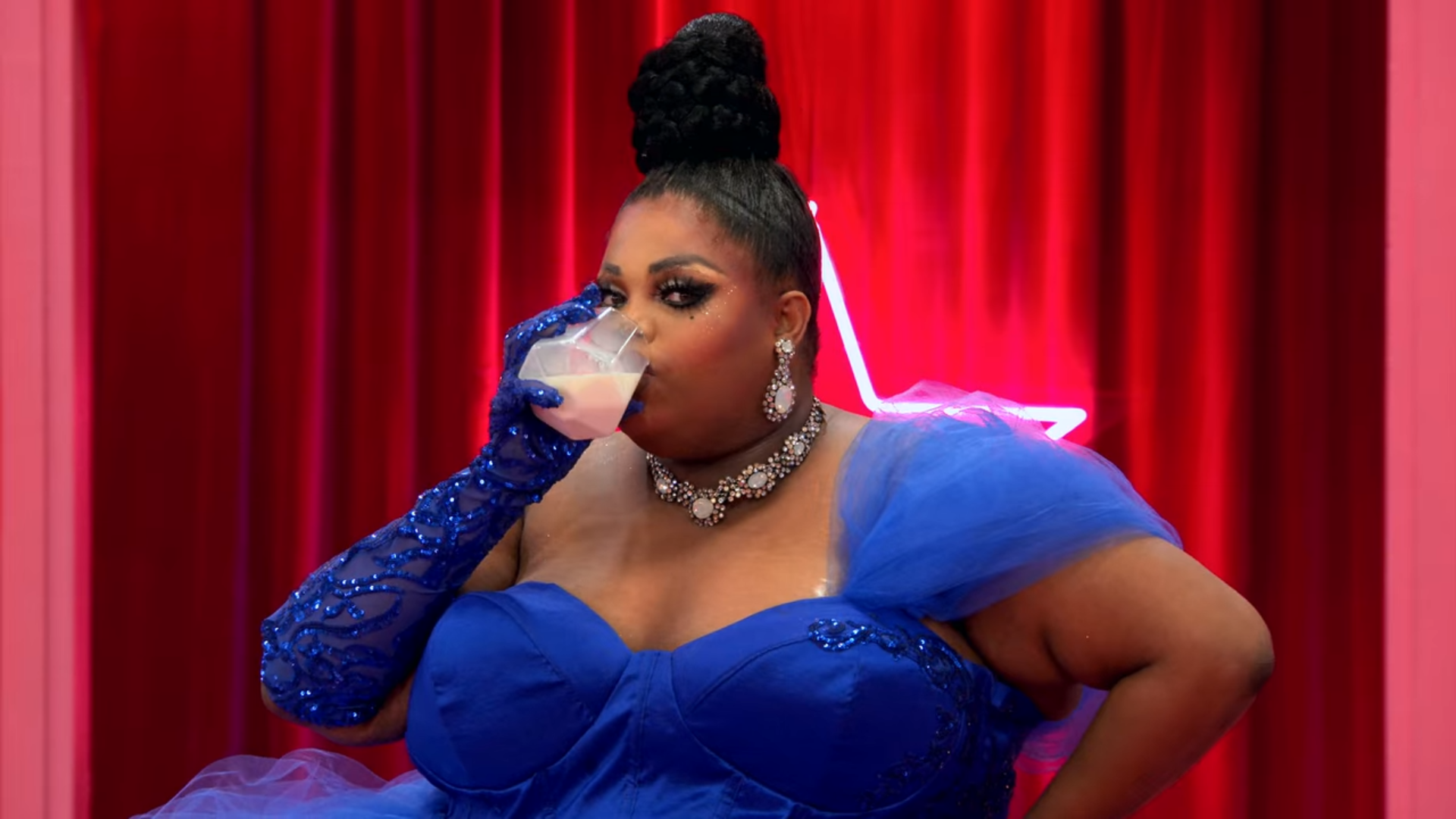 Cena do reality show All Stars 6, mostra a entrada de Silky Nutmeg Ganache, uma drag queen negra e gorda, que usa um vestido azul, rabo de cavalo alto e bebe um copo de leite, olhando para a câmara. Ao fundo, vemos uma estrela iluminada em um fundo vermelho.