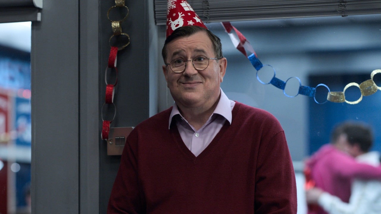 Cena da série Ted Lasso, mostra Higgins, um homem de meia idade e branco, sorrindo com um chapéu vermelho de festa na cabeça. Ele usa óculos e veste camisa social clara e um suéter vermelho por cima.