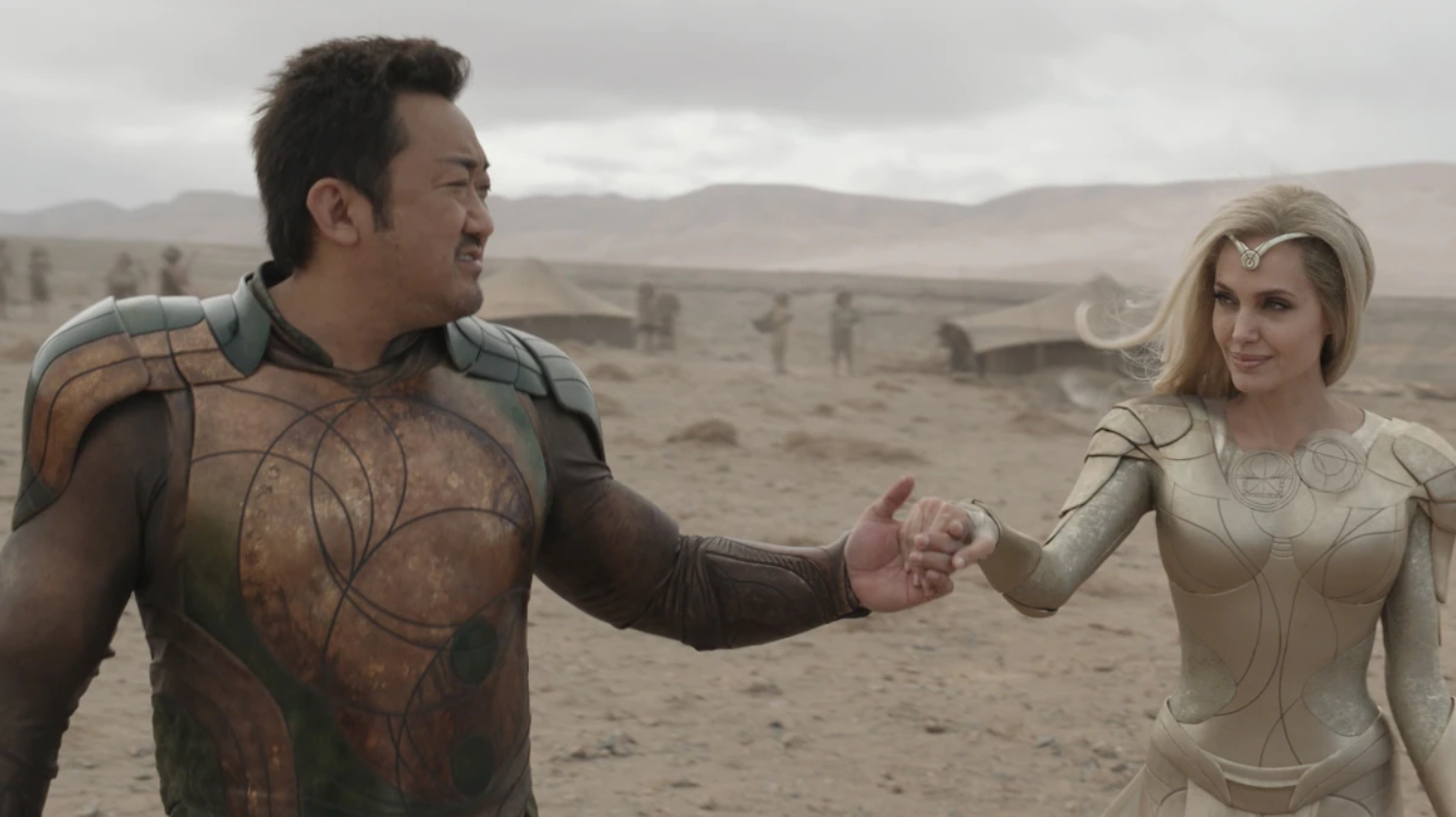 Cena do filme Eternos, mostra Gilgamesh dando as mãos para Thena em uma praia. Ele é um homem coreano e ela uma mulher branca.