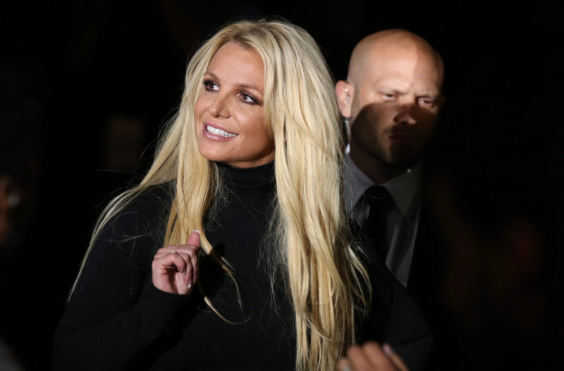 Foto de Britney Spears acompanhada do seu antigo chefe de segurança Edan Yemini. A cantora branca e loira veste preto enquanto sorri. Edan, um homem branco e careca, está posicionado atrás dela, ele usa uma camisa branca com terno e gravata pretos.