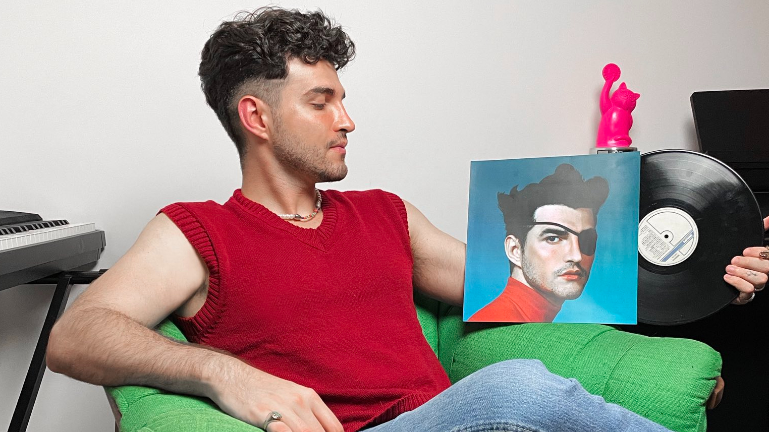 A imagem mostra o cantor Jão, um homem jovem e branco, sentado em uma poltrona verde. Ele está em sua casa, usando uma regata vermelha e calça jeans. Seu rosto está virado para a direita, direcionando seu olhar para o vinil segurado por sua mão.