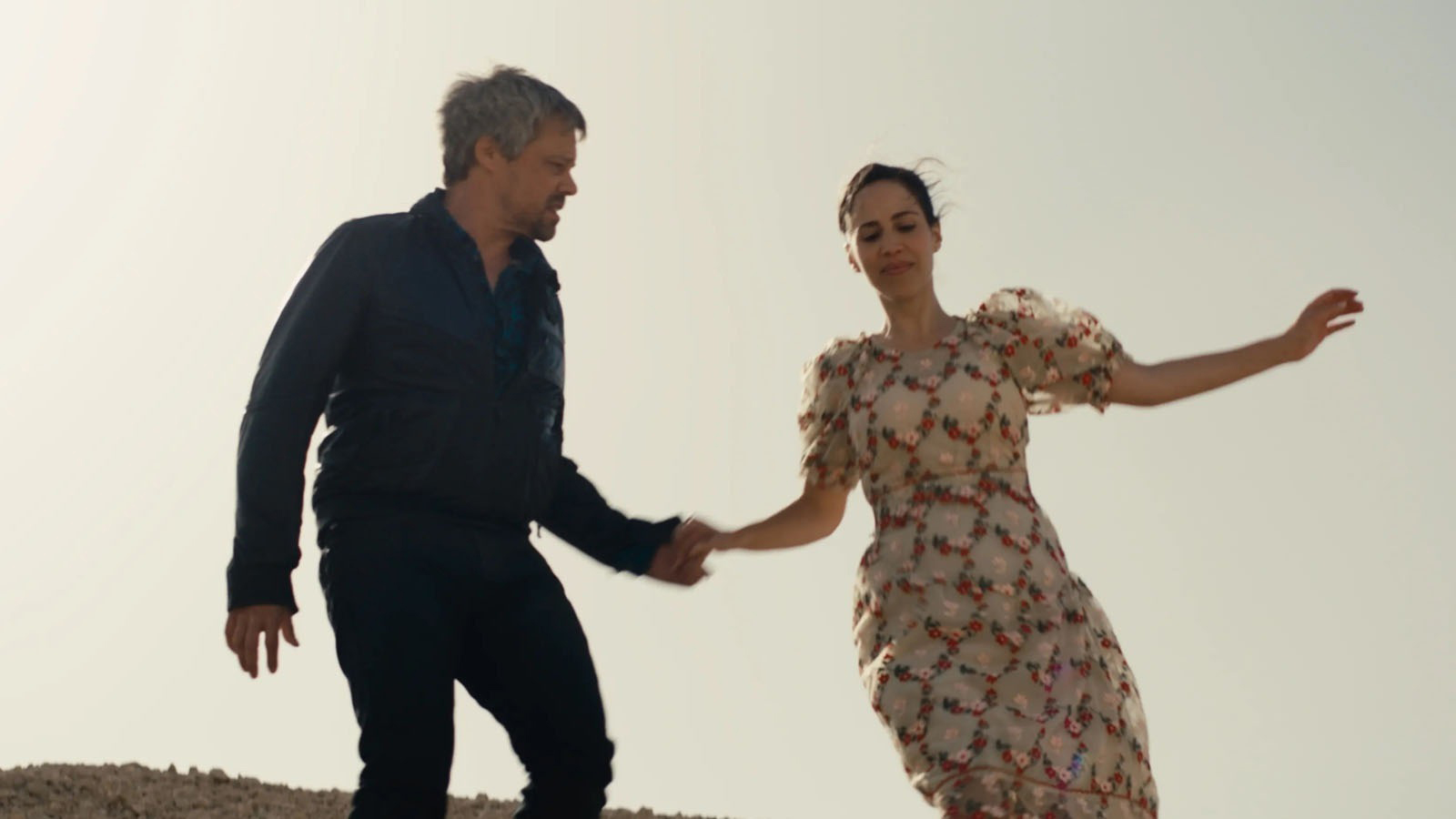 Cena do filme Ahed's Knee mostra um casal de mãos dadas, descendo uma colina. Ele usa roupas pretas e ela usa um vestido claro.