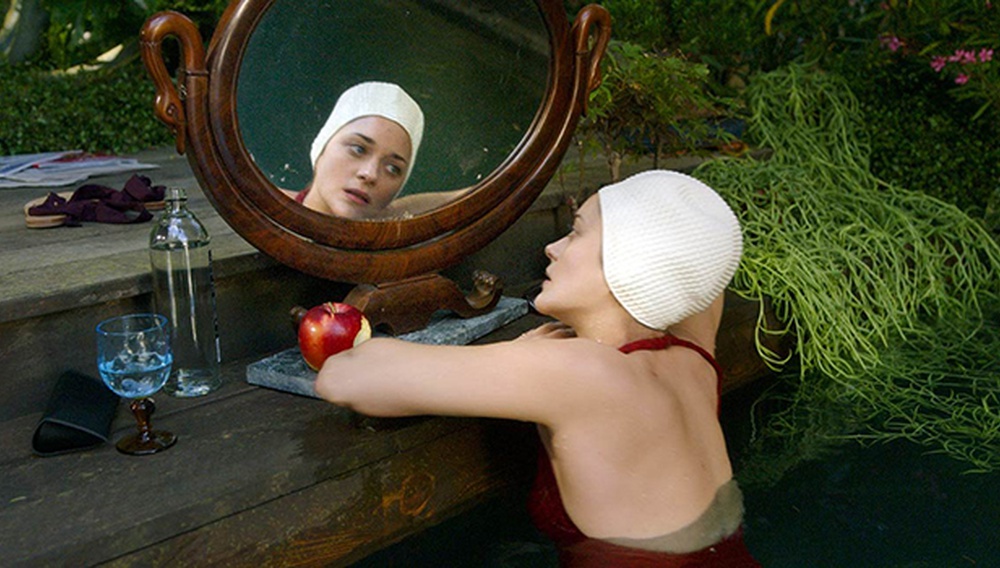 Cena do filme Annette exibe uma mulher branca olhando para um espelho enquanto está na piscina. Ela utiliza uma touca branca para cobrir o cabelo e um maiô vermelho. No degrau de madeira onde está apoiado o espelho, vemos uma maçã mordida, uma taça de vidro com água e uma garrafa d’água. 