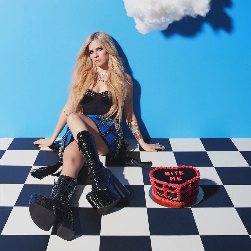 Capa do single Bite Me. No canto superior esquerdo, vemos as palavras “Avril Lavigne” em uma letra vermelha, em caixa alta, em uma fonte estilizada. No canto superior direito, vemos uma nuvem branca e a sombra. Em frente a uma parede azul clara, vemos a cantora Avril Lavigne ao centro, sentada em um chão quadriculado em preto e branco, como se fosse um xadrez. Avril Lavigne é uma mulher branca, de cabelos loiros lisos e compridos, vestindo uma blusa preta com brilhos brancos no busto, uma saia listrada azul e preta e botas pretas de couro de cano alto. Ela tem suas mãos apoiadas ao lado do corpo sentado e olha para a câmera. Ao lado direito dela, no chão, vemos um bolo preto em formato de coração com o contorno e as palavras “BITE ME” em vermelho. No canto inferior direito, vemos o símbolo do Parental Advisory.