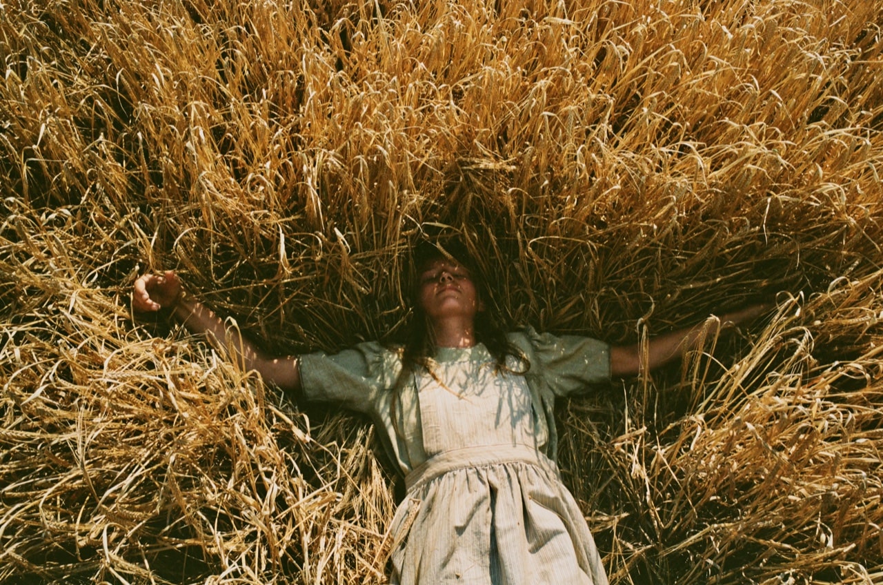 Cena do filme Assim Como no Céu. A imagem mostra uma jovem branca e loira deitada numa plantação alta amarelada. Ela usa um vestido verde, seus braços estão abertos e ela está de olhos fechados
