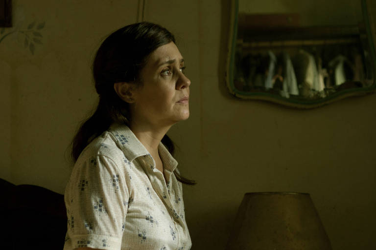 Cena do filme Marighella, mostra Adriana Esteves sentada e olhando para a janela. Ela é branca, tem cabelos escuros e é iluminada pela luz do sol que entra pela janela.