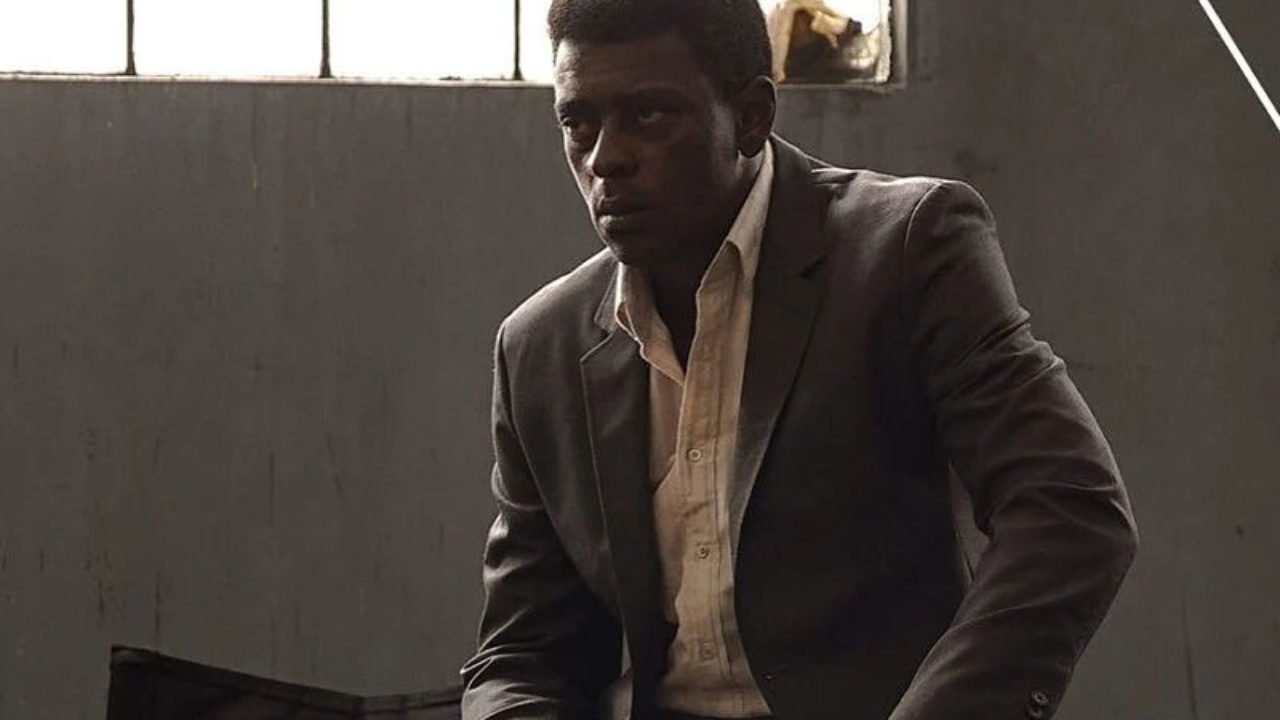 Cena do filme Marighella, mostra o protagonista Seu Jorge usando terno e sentado, olhando para a frente com a cara fechada. Ele é um homem adulto, de pele negra e cabelos pretos.