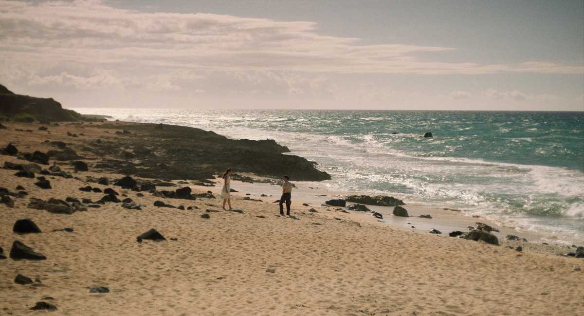 Cena do filme Eu Era um Homem Comum. A imagem mostra uma praia deserta com apenas duas pessoas nela. O dia está ensolarado. As pessoas são: uma mulher amarela e um homem amarelo. Eles vestem roupas da década de 1960.