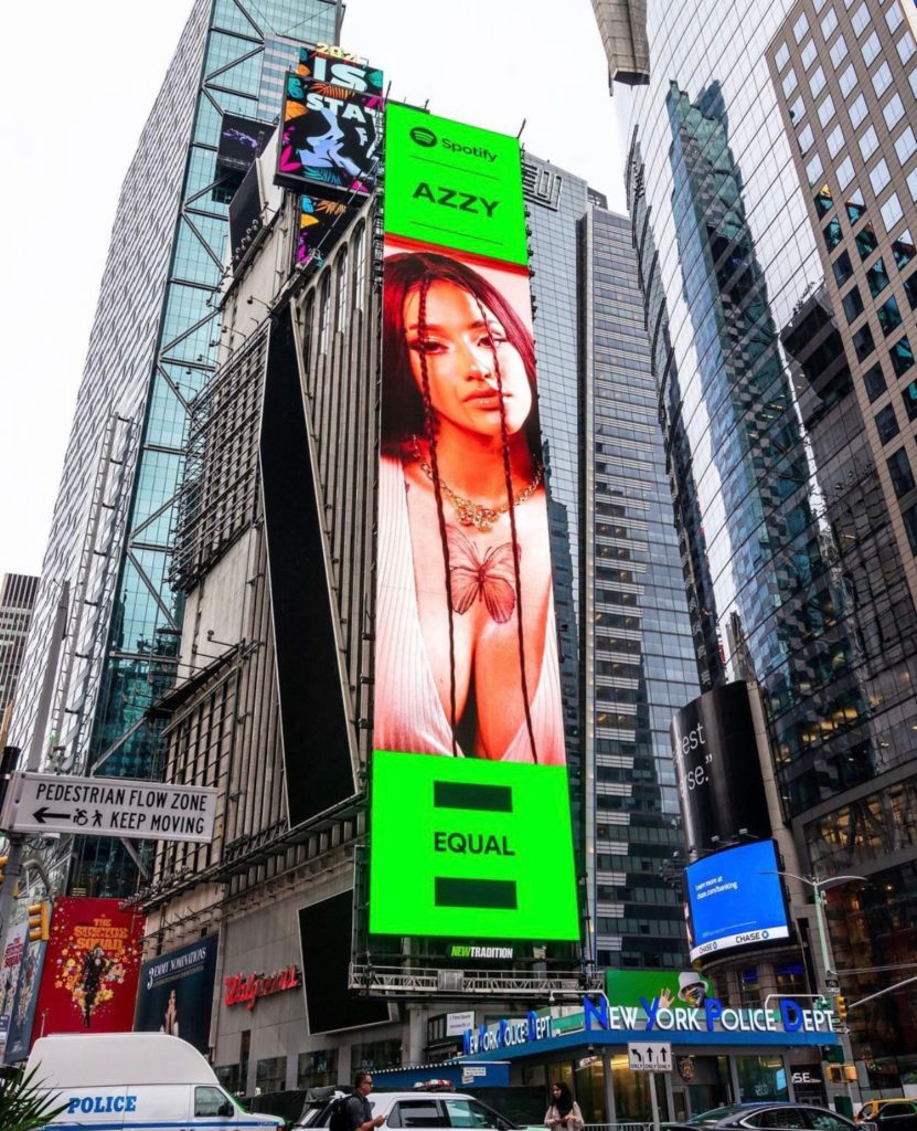 Imagem de um painel na Times Square, em Nova Iorque, que mostra Azzy como o rosto da campanha EQUAL do Spotify. A imagem é vertical e mostra vários prédios espelhados ao fundo. Ao centro, está o painel que estampa uma fotografia de Azzy.