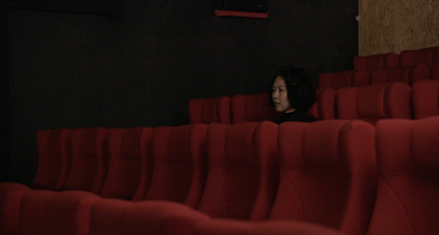 Cena do filme A Mulher que Fugiu. Gam-hee, interpretada por Kim Min-hee, é uma mulher coreana de cabelo curto preto com cerca de 39 anos. Ela veste uma blusa preta de gola alta e está sentada dentro de uma sala de cinema vazia. As poltronas da sala de cinema são vermelhas.