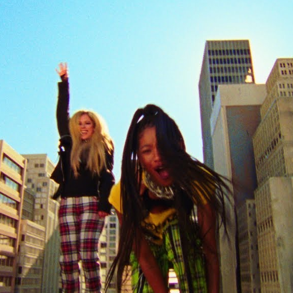 Cena do videoclipe de G R O W. Na imagem, à frente de um céu azul e de prédios nos cantos direito e esquerdo, vemos, ao centro, Avril Lavigne (à esquerda) e WILLOW (à direita). Avril Lavigne é uma mulher branca, de cabelos loiros lisos e longos, aparentando cerca de 30 anos, vestindo uma blusa preta, jaqueta de couro preta e calça listrada nas cores vermelha, preta e branca. Ela tem um de seus braços levantados, com a mão apontando para cima, e sorri. WILLOW é uma mulher negra, de cabelos pretos longos, aparentando ter cerca de 20 anos, usando um calor prateado, vestindo uma blusa amarela e calça listrada nas cores verde e preto. Ela está inclinada em direção à câmera com a boca aberta, no que aparenta estar cantando.