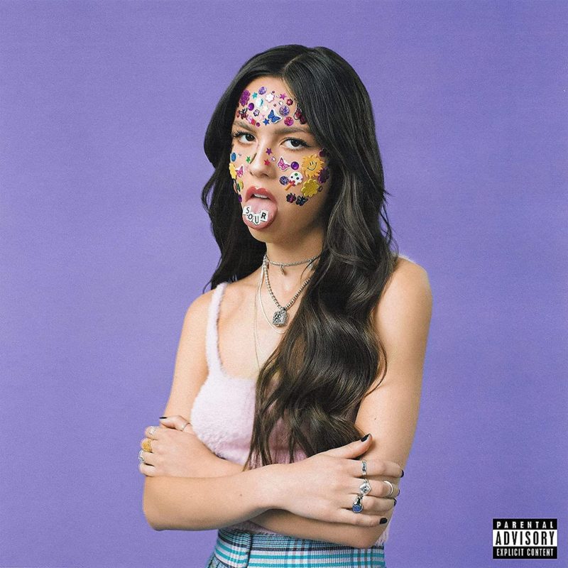 Capa do álbum SOUR. Mostra Olivia Rodrigo, mulher jovem, magra e de origem filipina, com a língua de fora e os olhos revirados. Ela tem vários adesivos colados no rosto e o fundo da imagem é roxo. 