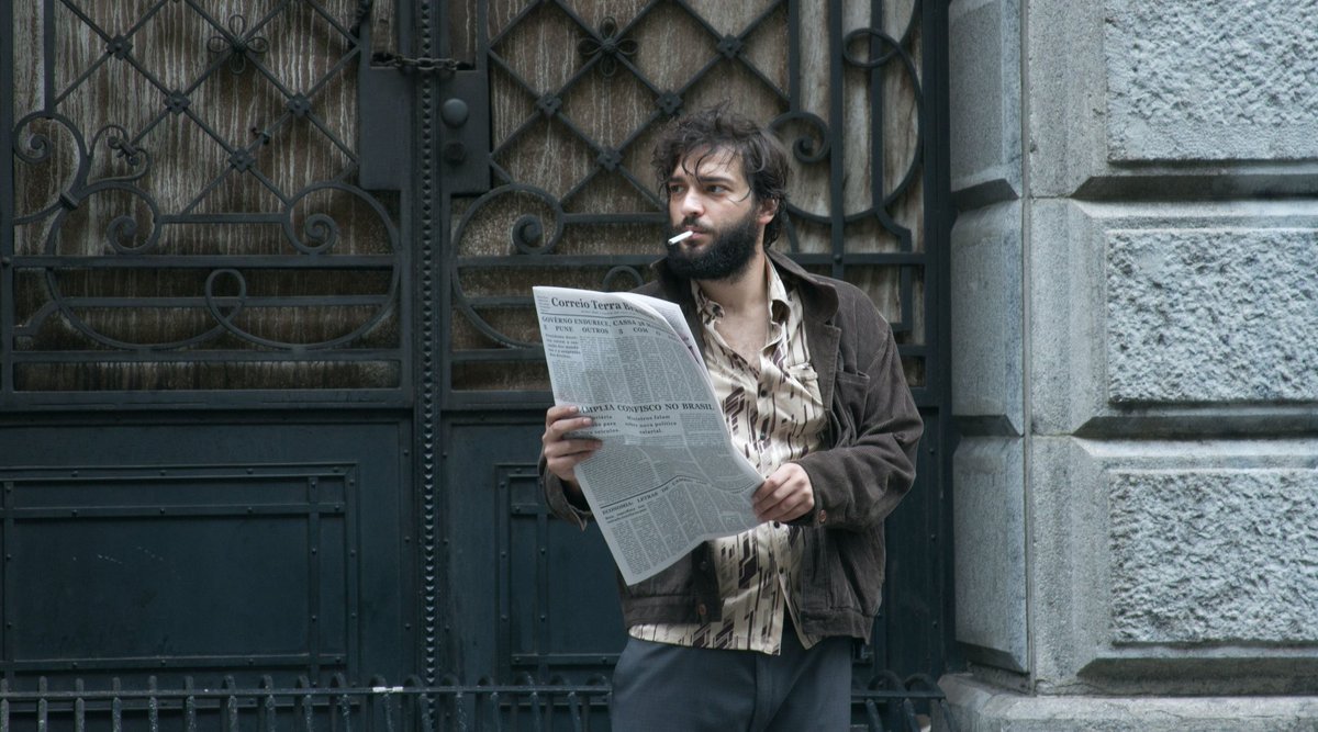 Cena do filme Marighella, mostra o ator Humberto Carrão com um jornal na mão e fumando na calçada. Ele é branco, tem barbas e cabelos volumosos e escuros e usa uma camisa estampada marrom com o botão de cima aberto.