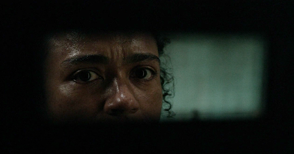 Cena da série The Walking Dead, mostra uma fresta escura, iluminada no meio e que mostra os olhos de Connie, uma mulher negra que está suada e com expressão de cansaço.