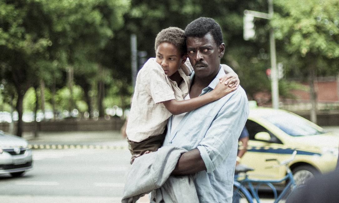 Cena do filme Marighella, mostra Seu Jorge, um homem negro e adulto, com o filho no colo, uma criança que usa camisa branca e se abraça ao pescoço do pai. A cena é de dia e ao fundo vemos árvores e carros.