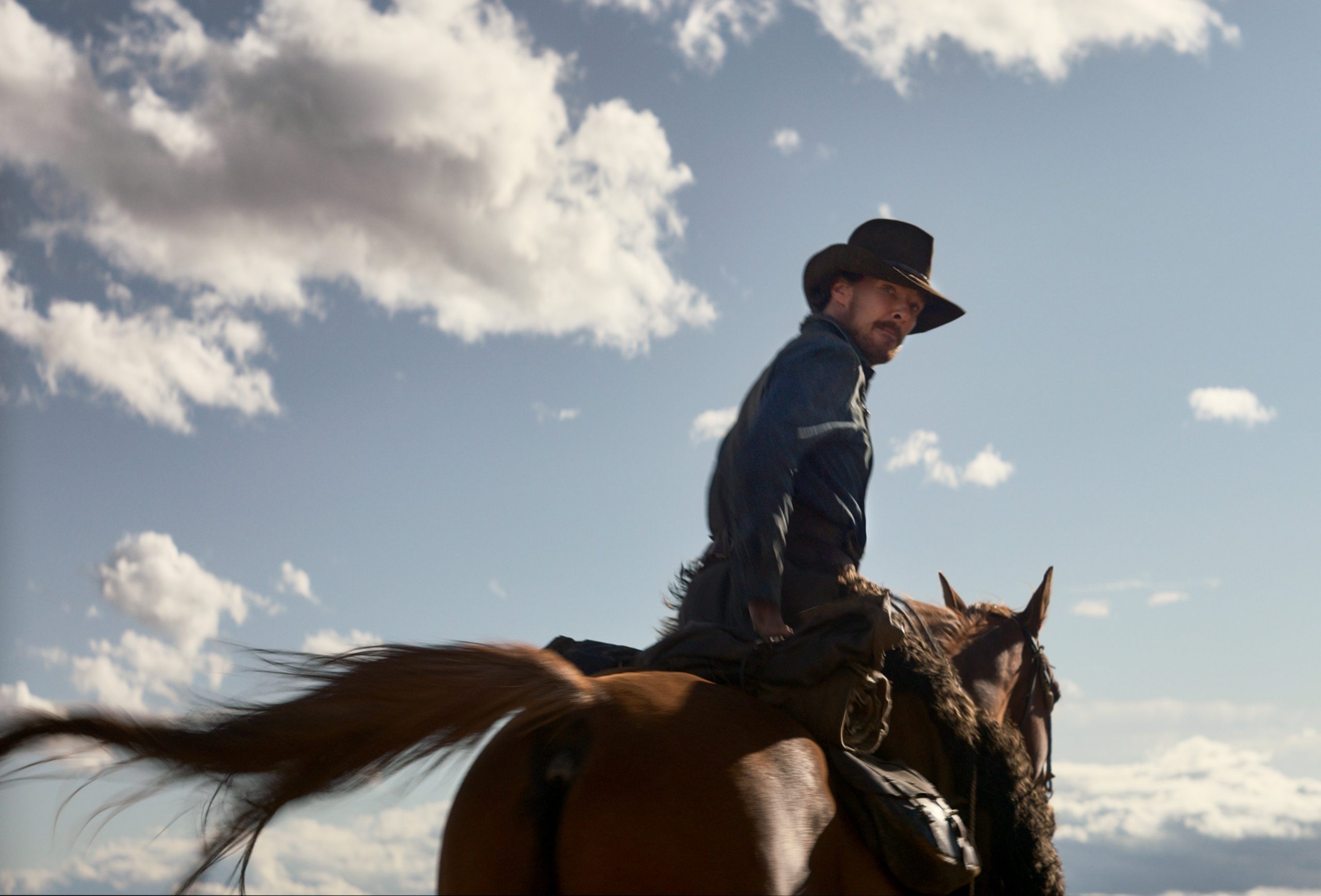 Cena do filme Ataque dos Cães, mostra o ângulo de trás de Phil montando em um cavalo. Ele usa roupas de cowboy e olha para baixo, montado no animal marrom. Na frente deles, vemos o céu azul cheio de nuvens.