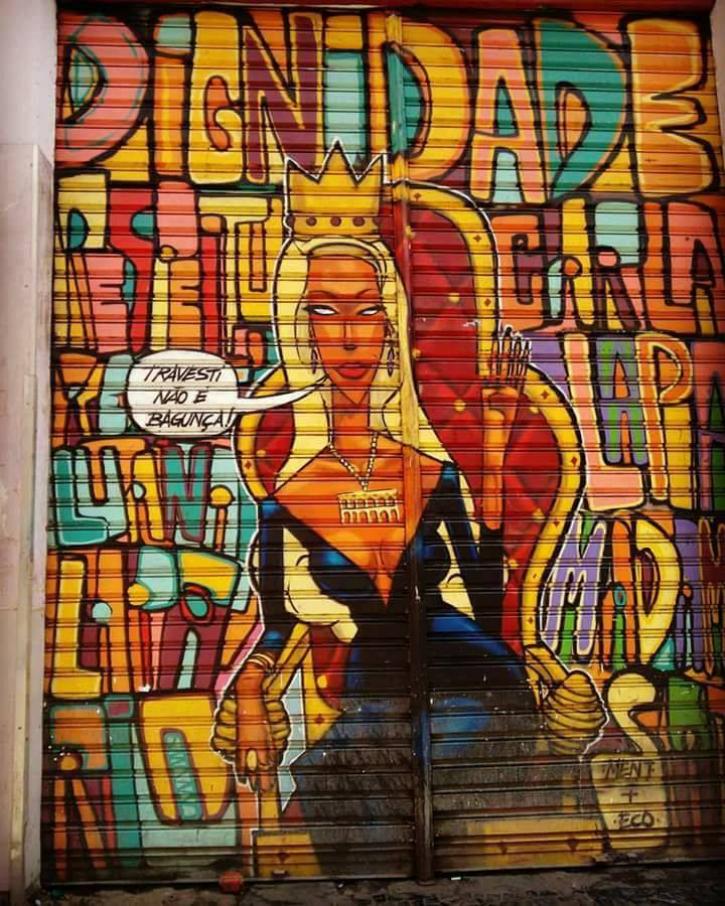Foto vertical de um grafite. Nele, vemos a representação de Luana Muniz com uma coroa, vestido preto, sentada no trono e com um balão de fala "TRAVESTI NÃO É BAGUNÇA". Ao redor, várias palavras completam o desenho como "LAPA" "DIGNIDADE" e "RESPEITO"