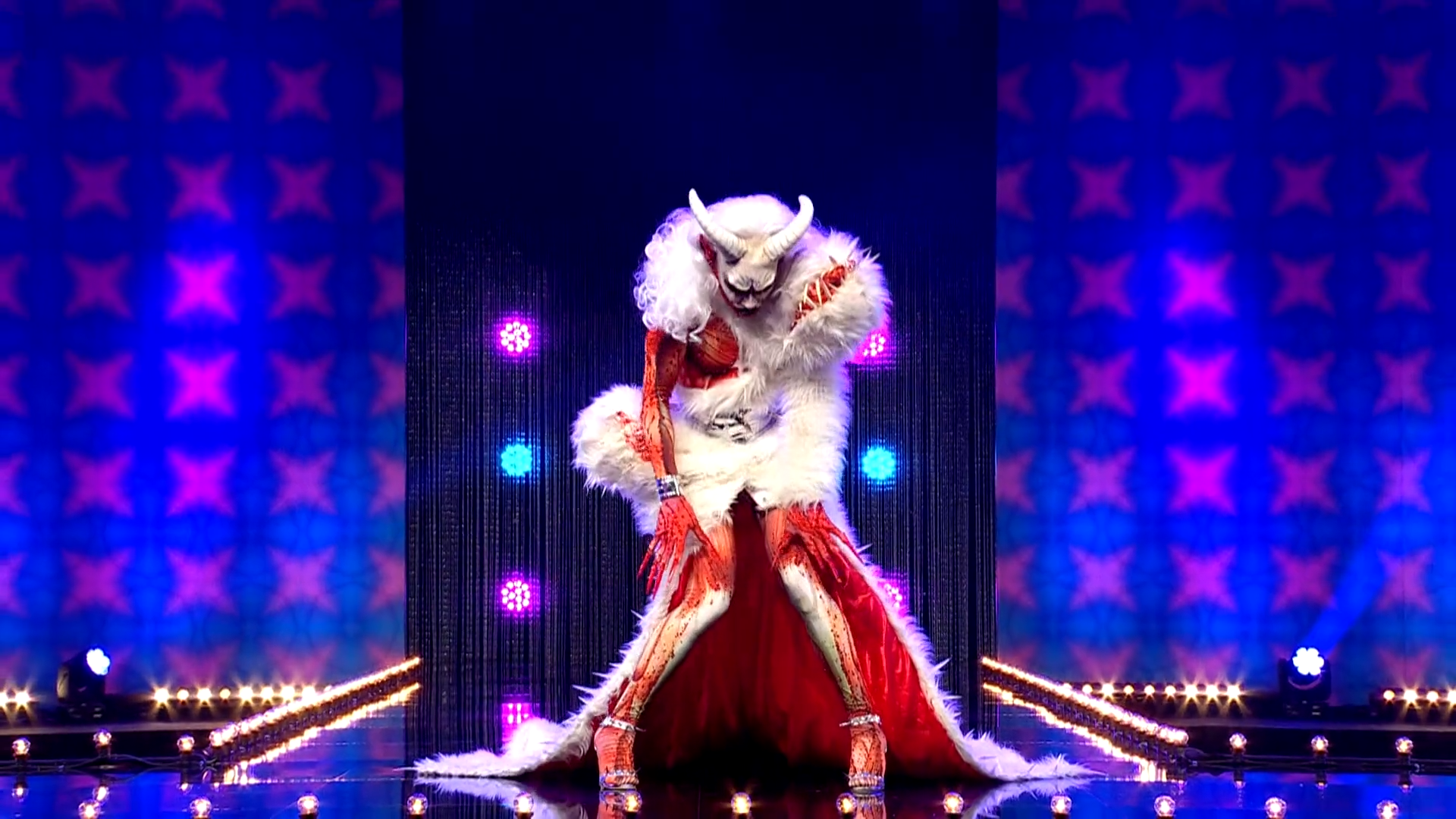 Cena de Drag Race Holland, mostra a drag Keta Minaj abaixada na passarela, usando uma roupa bestial, com ilusão de carne viva na pele e uma cobertura monstruosa e branca, como uma fera, com chifres brancos.