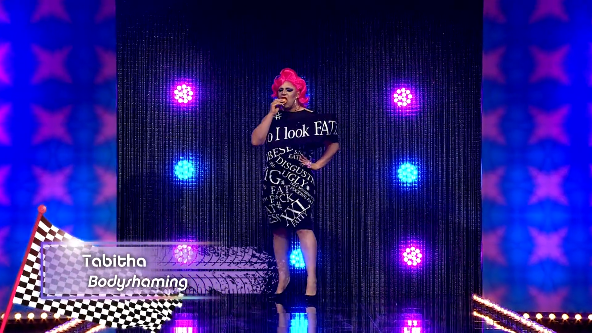 Cena de Drag Race Holland, mostra a drag queen Tabitha na passarela. Ele é branca, gorda, usa peruca rosa e vestido preto com escritas em branco. Ela come alguma coisa, com o alimento sendo mordido. No canto inferior esquerdo, vemos uma legenda escrito Tabitha e Bodyshaming.