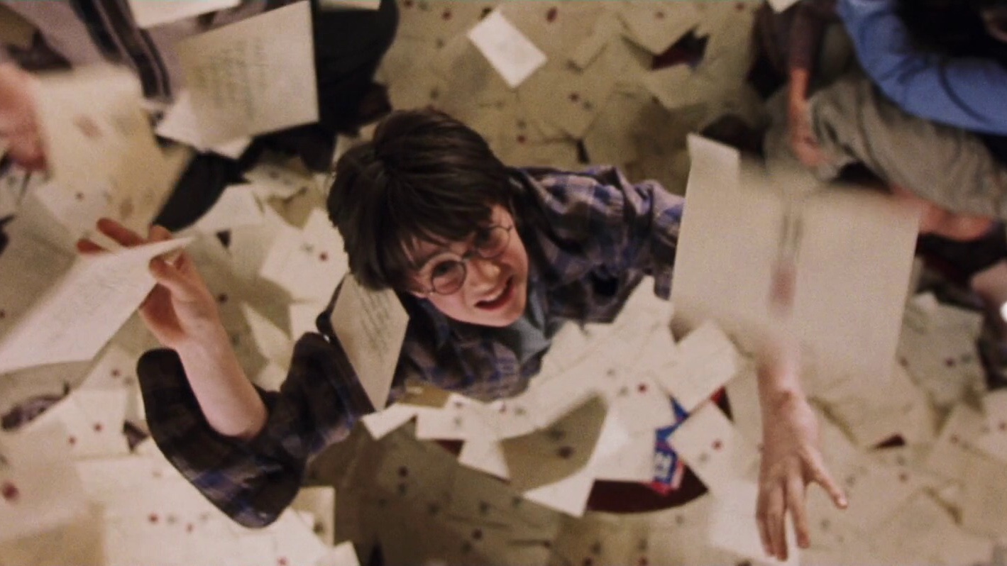 Cena do filme Harry Potter e a Pedra Filosofal mostra um jovem branco, de cabelos pretos e óculos redondo, que veste uma jaqueta azul, com as mão elevadas tentando pegar uma das cartas ao seu redor. O fundo da imagem é coberto com mais cartas.