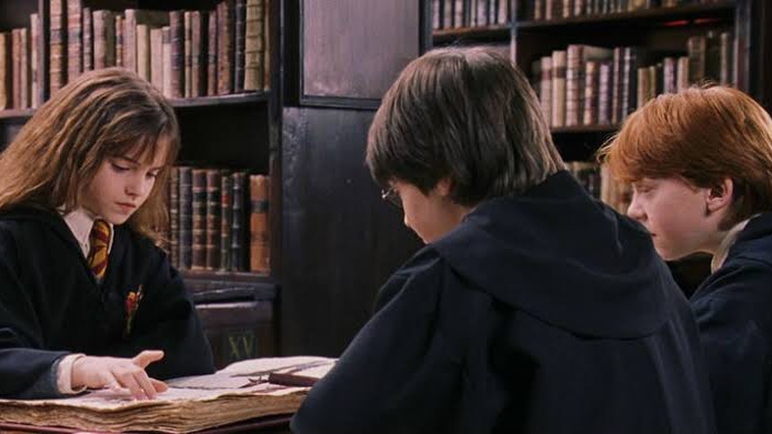 Cena do filme Harry Potter e a Pedra Filosofal apresenta três jovens sentados em uma biblioteca. À esquerda está uma menina branca, de cabelos castanhos claros armados, vestindo o uniforme de Hogwarts e lendo um livro. No meio está um menino branco de cabelos pretos, de costas para a câmera, vestindo o uniforme de Hogwarts. A direita está um menino branco e ruivo de perfil para a foto, vestindo o uniforme de Hogwarts.