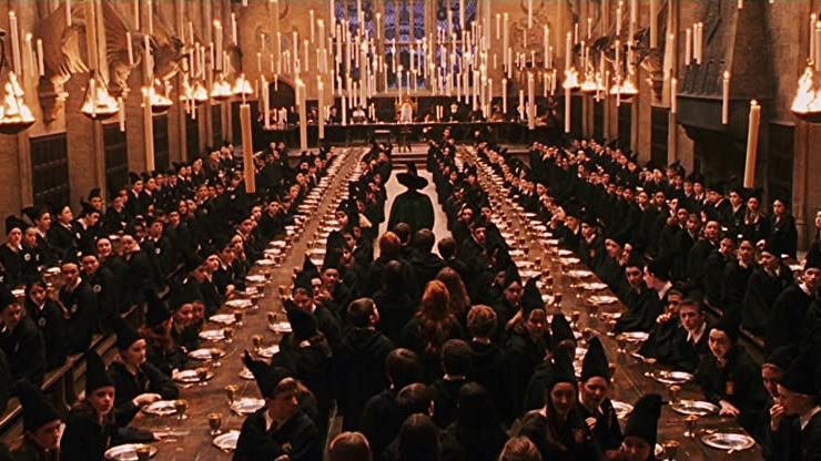 Cena do filme Harry Potter e a Pedra Filosofal apresenta todos os alunos uniformizados entrando no salão principal de Hogwarts, guiados por uma professora que usa um chapéu grande e pontudo. O local apresenta quatro mesas compridas com alunos de outros anos já sentados e velas flutuantes por todo o ambiente.