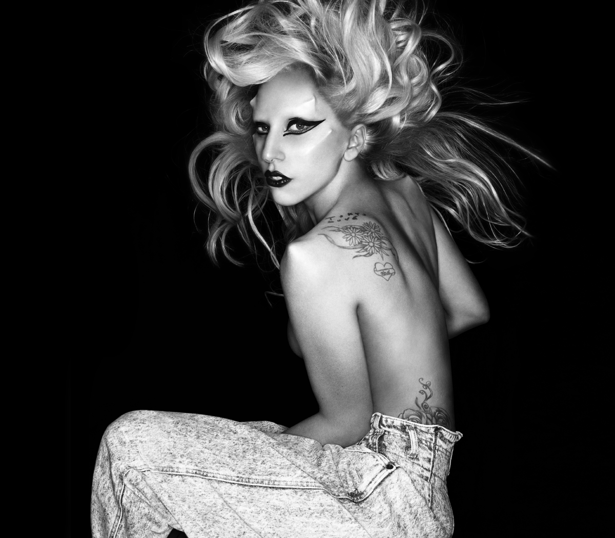Fotografia em preto e branco. A imagem mostra a cantora Lady Gaga de lado, sem sutiã, mas escondendo os seios com o próprio corpo. Ela está com os cabelos para cima, maquiagem com delineador, e usa calça. Ela olha diretamente para a câmera e tem próteses no rosto, imitando chifres.