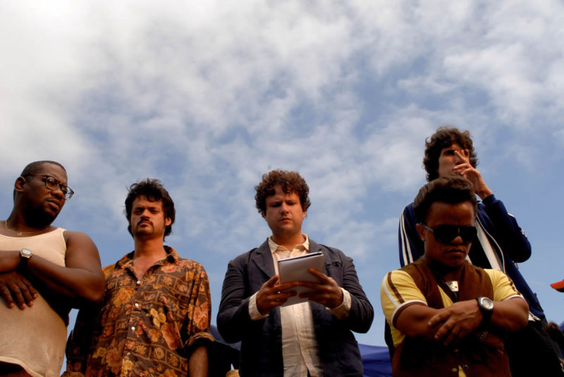 A imagem é uma cena do filme O Palhaço. Nela, estão, da esquerda para a direita, os atores Thogun, Álamo Facó, Selton Mello e Renato Macedo, com o ator Tony em frente a este último. 