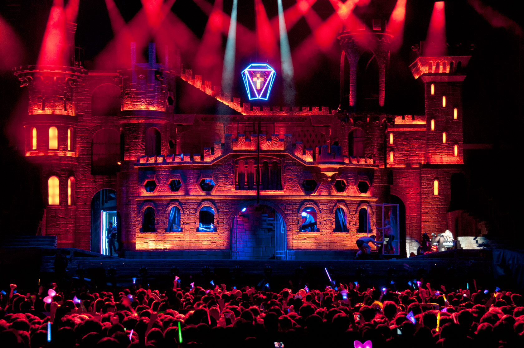 Fotografia retangular colorida. A imagem mostra o palco da Born This Way Ball Tour, um castelo medieval. Ele está iluminado por luzes vermelhas alaranjadas.